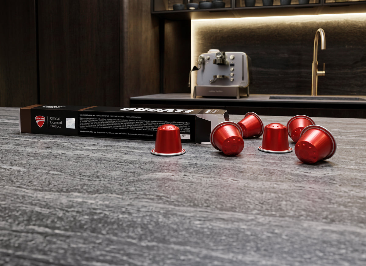 Caja de cápsulas Espresso by Ducati y cápsulas rojas sobre una encimera, con la Cuisine Barista M3 al fondo